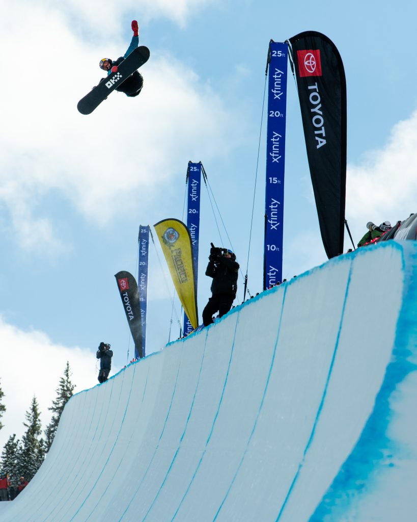 a snowboarder does a trick above the edge of a halfpipe, large banners with brand logos in the background