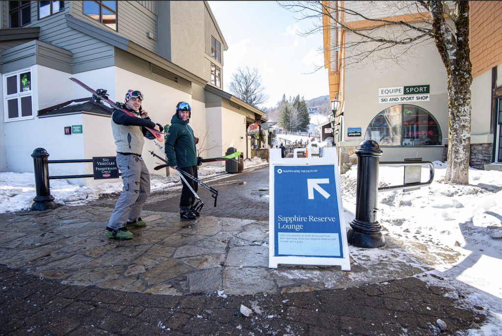 two skiers walk past a sign directing people to a lounge space