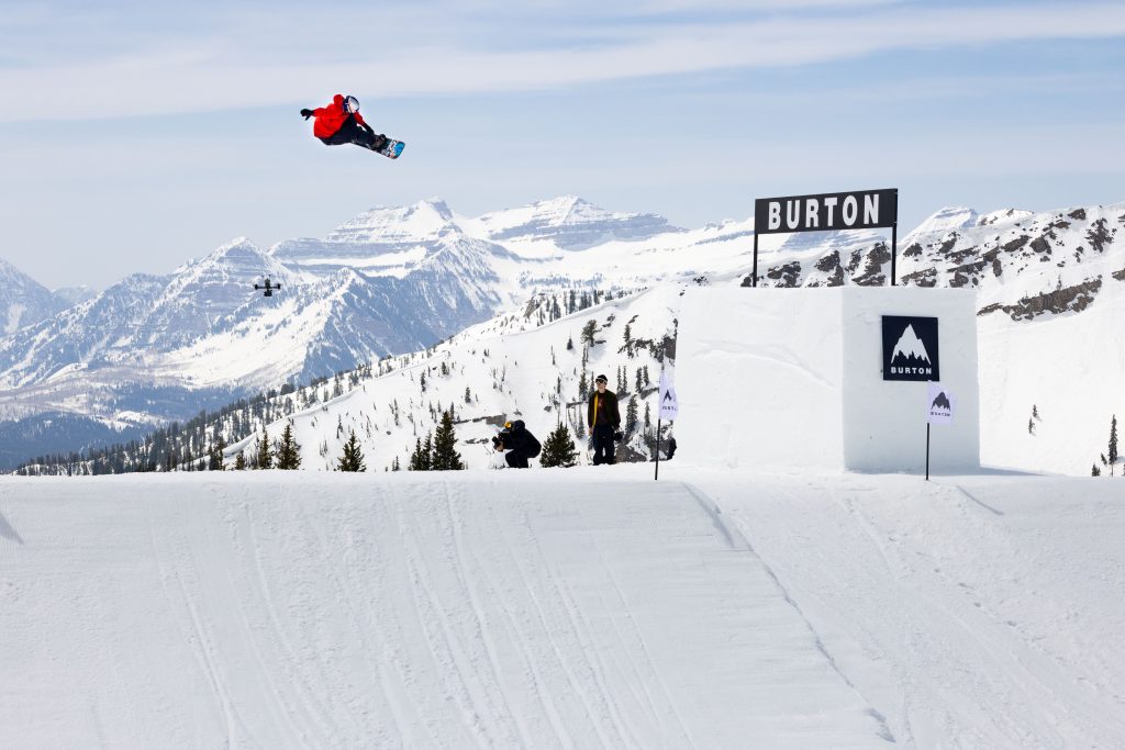 Jake Canter hits a jump on his snowboard, mountains in the background, burton branding on banners alongside the course