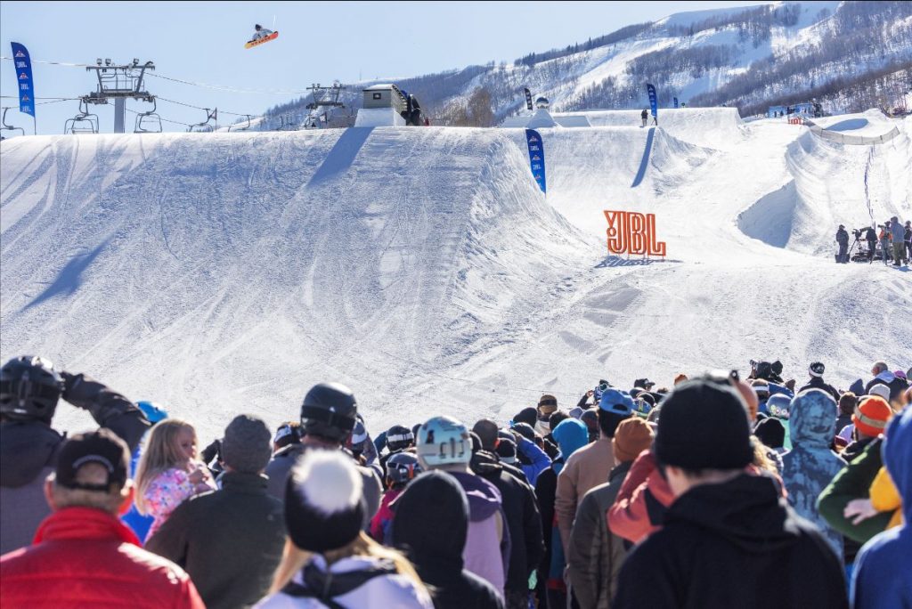 a snowboarder goes off of a jump, a crowd watches in the foreground