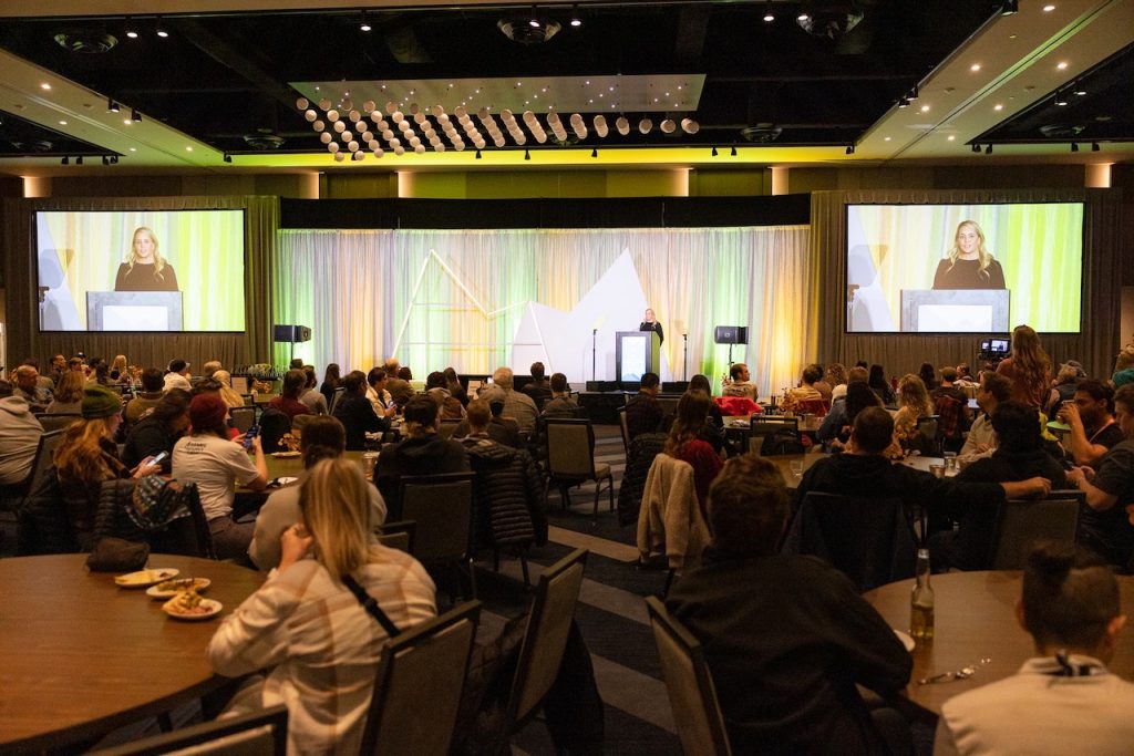 a speaker stands on a stage, a crowd in a banquet hall watches