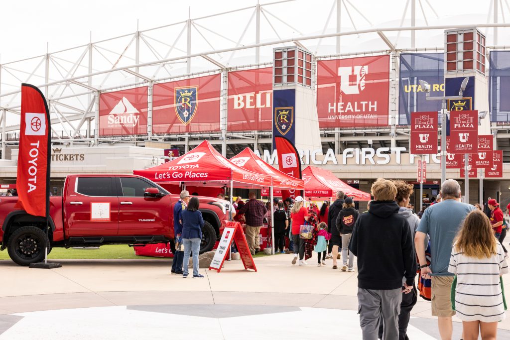 a crowd of people approach a stadium, walking past toyota-branded tents and truck