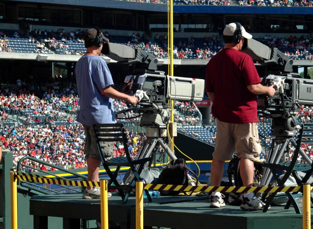 two camera operators in a stadium look through viewfinders at the field